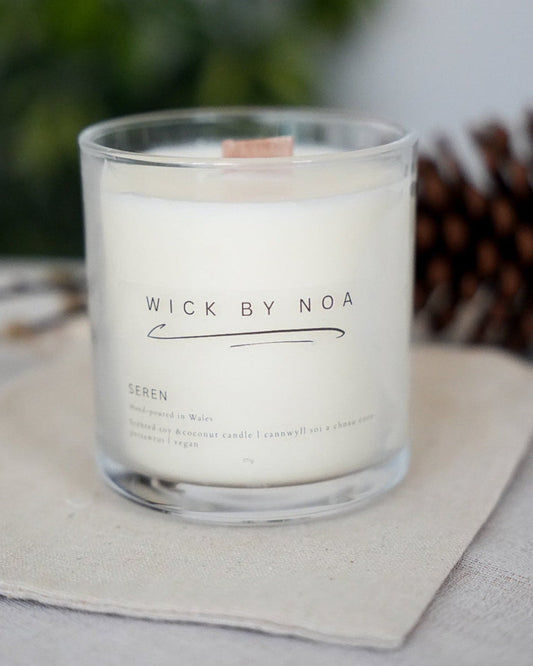 Candle in a glass jar labeled 'Wick by Noa' on a light surface with a blurred natural background